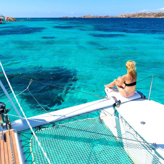 Woman in bikini tanning and relaxing on a summer sailin cruise, sitting on a luxury catamaran in picture perfect turquoise blue lagoon near Spargi island in Maddalena Archipelago, Sardinia, Italy.